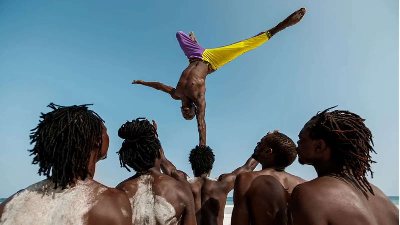acrobat on the beach, balancing upside down with his hand on another acrobat's head; four others watch