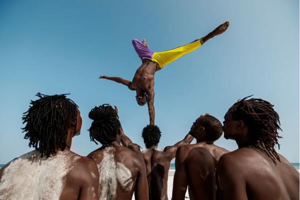 acrobat on the beach, balancing upside down with his hand on another acrobat's head; four others watch