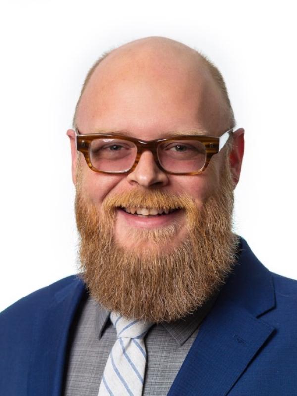 A photo of Bennett Whitaker, a smiling white-presenting person with very short blond hair, a mustache and a medium length beard. Bennett is wearing brown eyeglasses, a bright blue suit coat, a subtle gray plaid shirt and a white tie with narrow blue stripes.