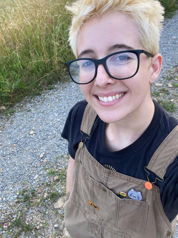 Selfie taken by Daisy Ahlstone smiling with bleach blonde hair, black glasses, and wearing light brown overalls and a black t-shirt, outdoors on a gravel path with grass in the background.