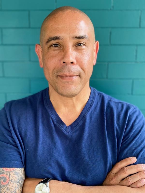 standing with arms crossed in a blue shirt against a teal brick background