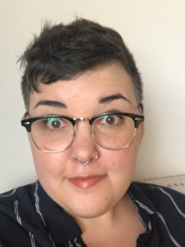 A short-haired nonbinary white person (brown-haired) with browline glasses and a happy expression on their face. They are wearing a blue shirt with white stripes.