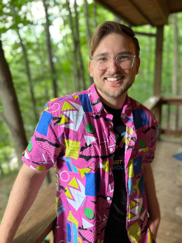 A man with clear glasses; pink, 90s-themed, open button-down shirt; smiling in the woods