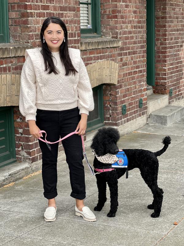 A photo of Dr. Maya Cruz smiling in a white top and black pants, standing alongside her service dog Rosalie, a black standard poodle in a blue vest.