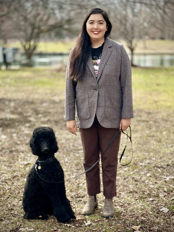 Maya is standing in a brown suit next to her Black standard poodle who is sitting. They are both staring at the camera straight on and Maya is smiling.