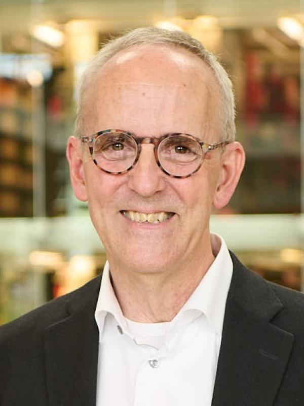 Head and shoulders of man with short gray hair and glasses in front of library bookshelves